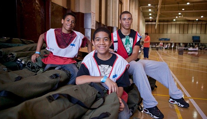 Three young male volunteers wearing Red Cross vests sitting on cots in a gymnasium