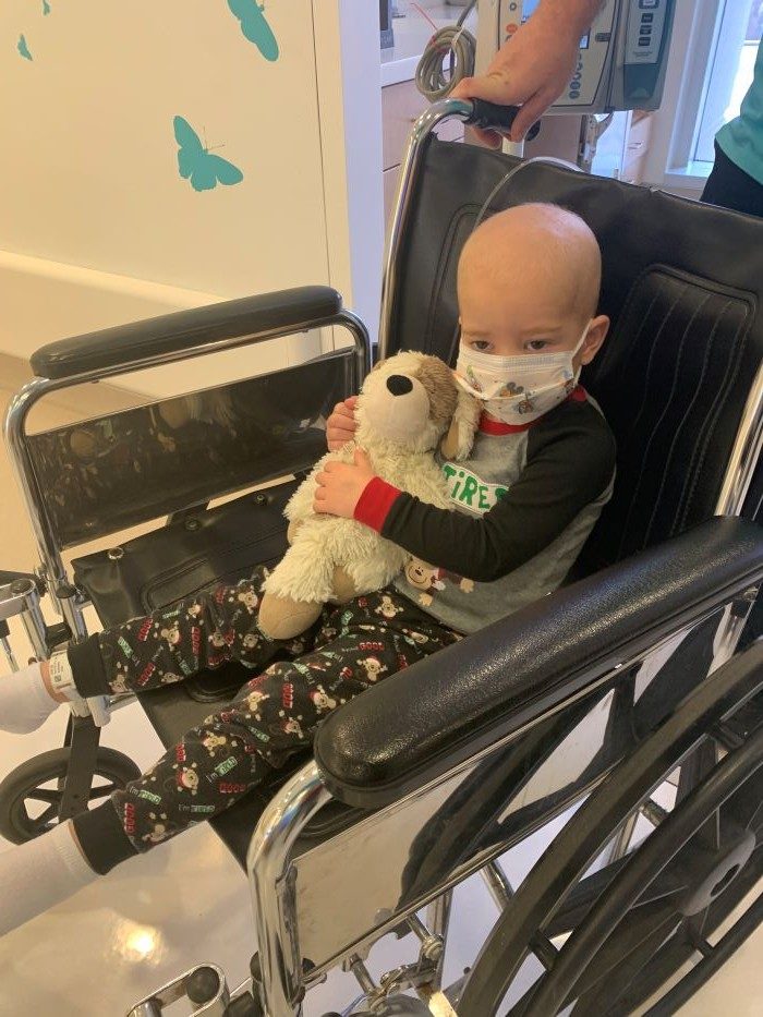 Toddler Cole Kulman in awheelchair wearing pajamas and a face mask holding a teddy bear