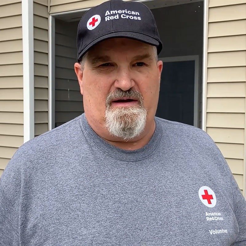 Bill Schmidt in front of house wearing Red Cross hat and shirt