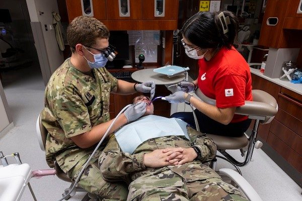 A young woman participates in chairside training at a military dental clinic. She’s helping a uniformed dentist with a tooth cleaning.