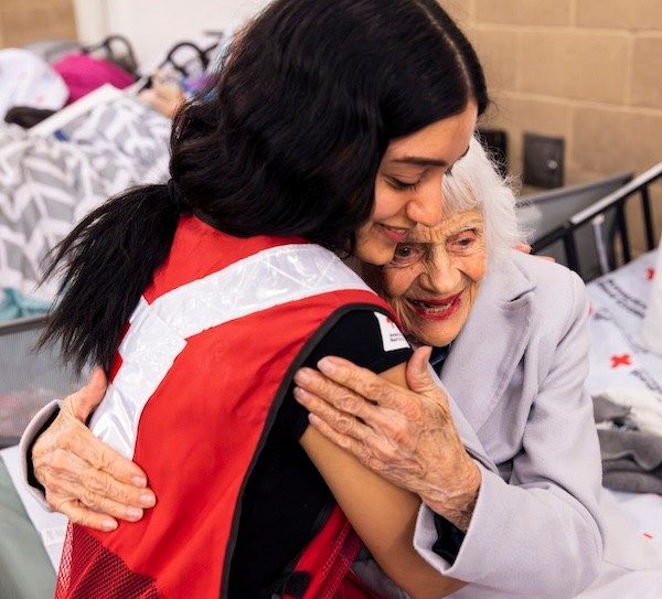 A young Red Cross volunteer with long black hair wearing a red vest hugs an older woman at a shelter.