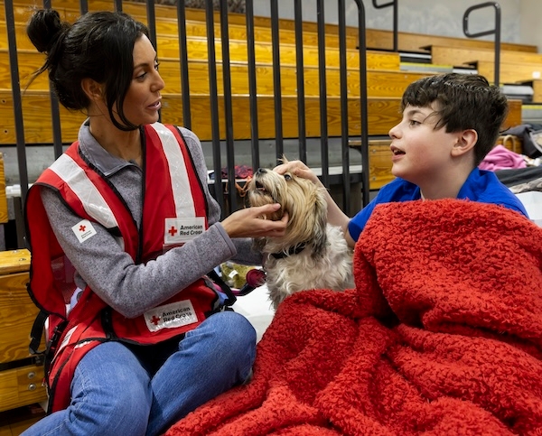 A Red Cross volunteer in a vest pets a a dog while sitting with a young boy in a shelter.