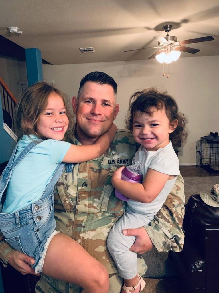 man in military fatigues holds his two daughters and smiles at the camera