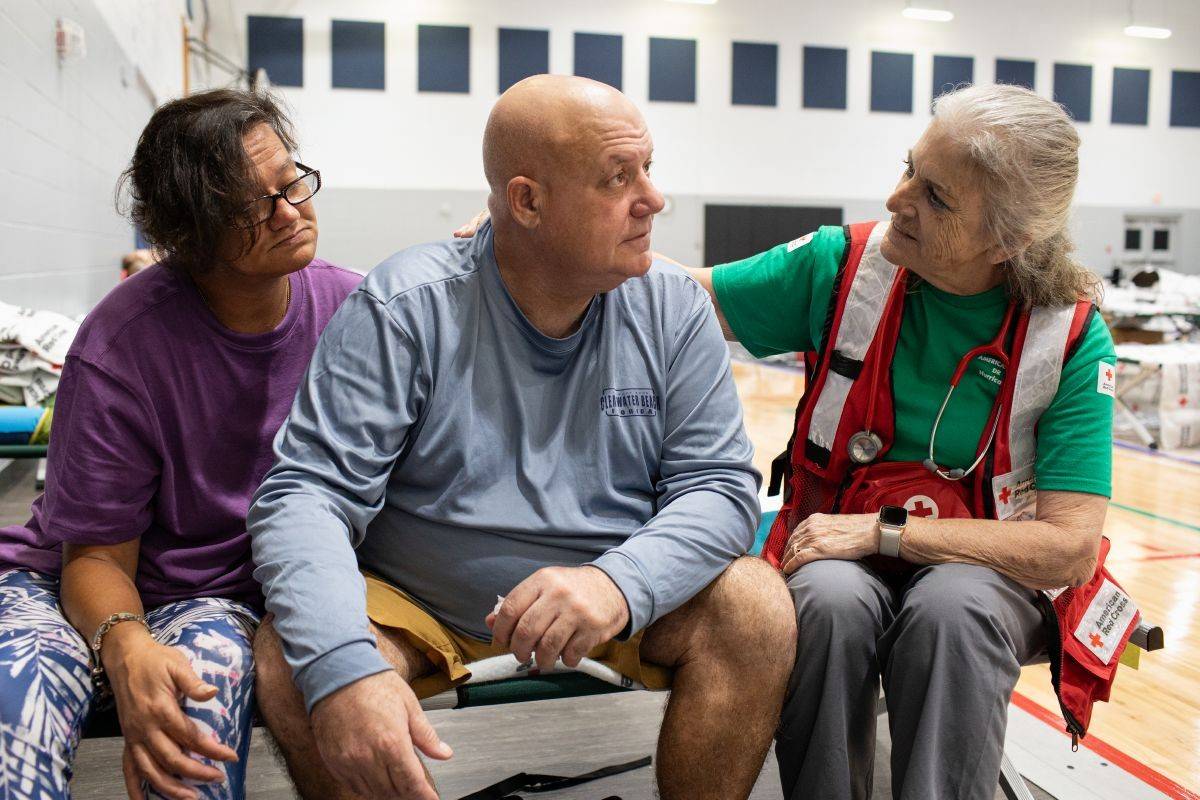 Un voluntario de Cruz Roja está sentado en un catre ayudando a dos personas que se encuentran alojadas en un refugio.