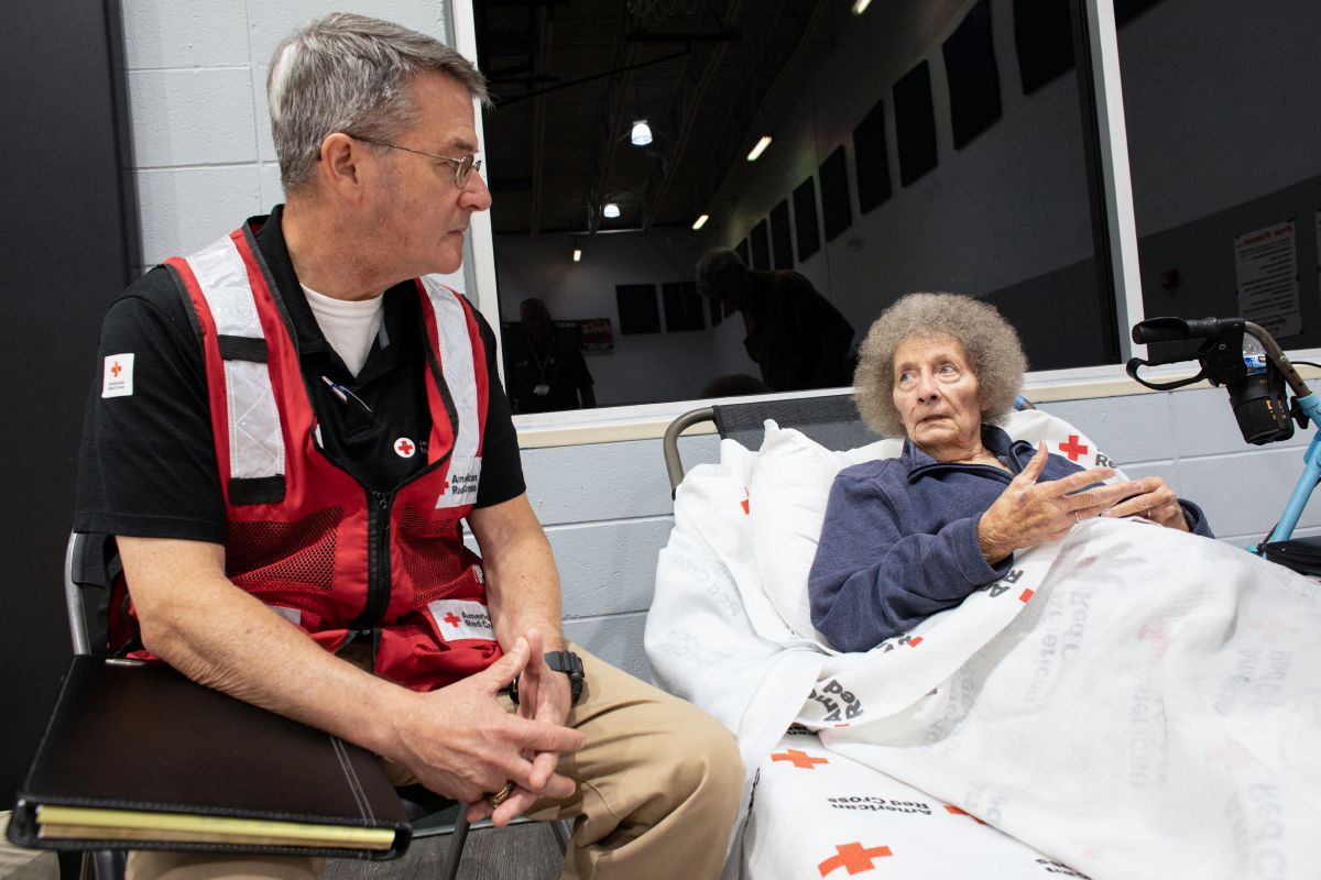 Red Cross volunteer sitting next to person who is laying in cot with a Red Cross blanket.