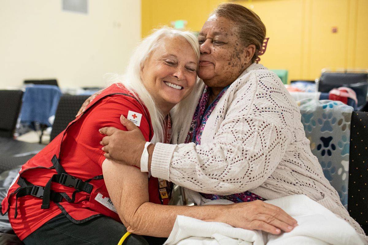 A Red Cross volunteer is sitting on a cot in a shelter hugging a hurricane survivor, they are both smiling.