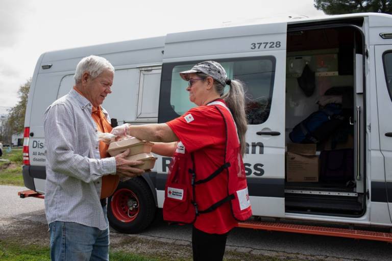 Woman in Red Cross vest hands meals to men