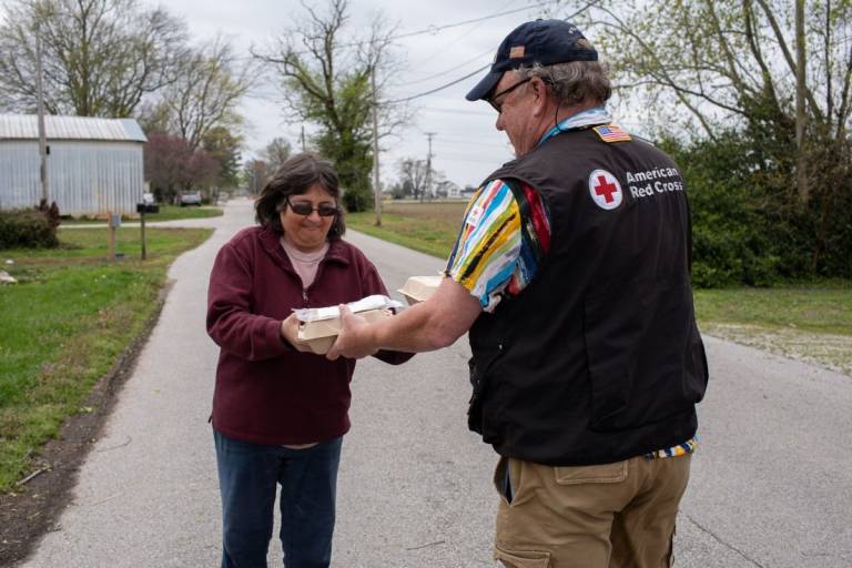 Man in Red Cross vest hands meals to woman