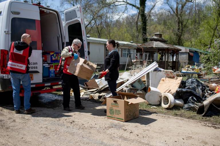 Man in Red Cross vest delivers essential supplies to woman in front of piles of flood debris