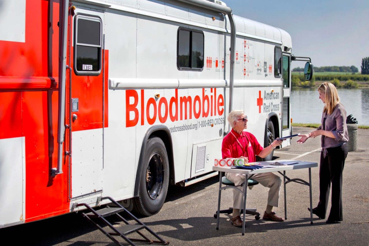 Red Cross bloodmobile vehicle with Red Cross volunteer sitting at table next to it and handing pen to woman.