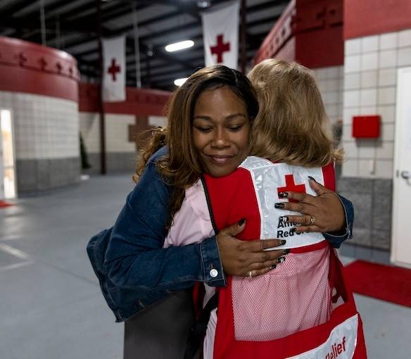 A Red Cross Volunteer embraces a woman standing in the hallway of a building with Red Cross banners hanging from the ceiling.