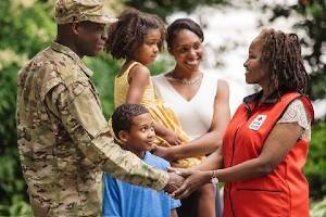 An American Red Cross volunteer smiling and holding the hand of an armed forces man with his family.