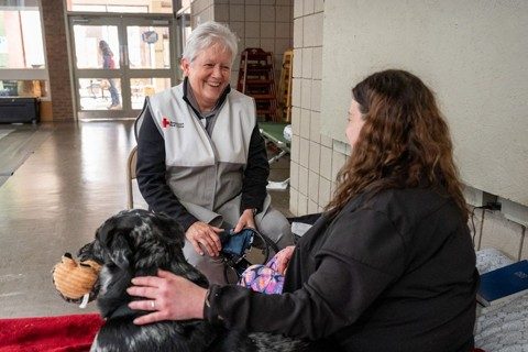 A Red Cross nurse is checkin on a woman in a Red Cross shelter. The woman is sitting on a cot with her dog.