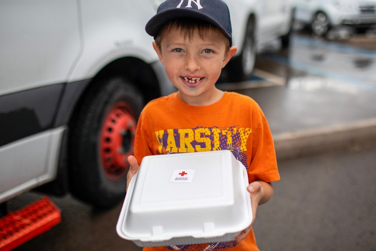 A smiling young boy in an orange shirt holds a food container with a sticker of the Red Cross logo