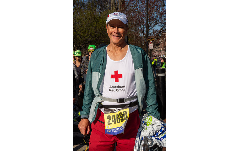 Marathon runner smiling for camera wearing hat, jacket, Red Cross shirt and marathon number on waist