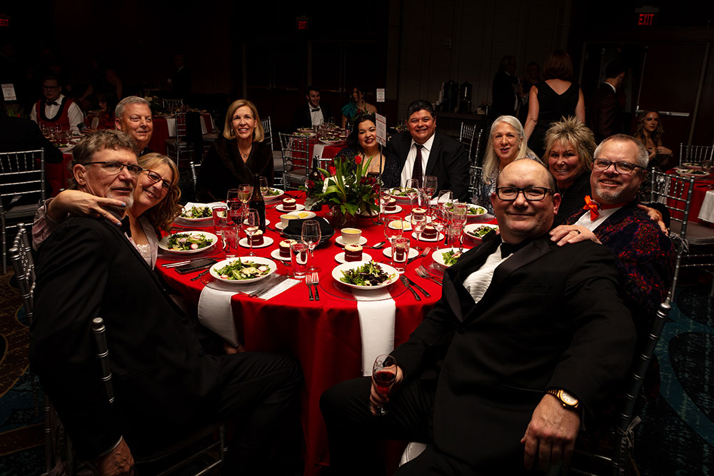 Red Cross Gala guests sitting at a round table with plates of food and drinks and smiling for the camera.