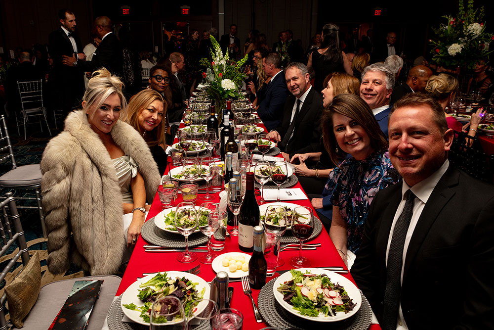 Red Cross Gala guests sitting at a long rectangle table with plates of food and drinks and smiling for the camera.