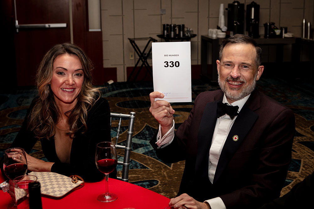 Two people at Red Cross Gala, sitting at table with one person holding a piece of paper up that has a number on it.