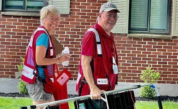 Smiling man and woman in Red Cross vests carrying a ladder and clipboard