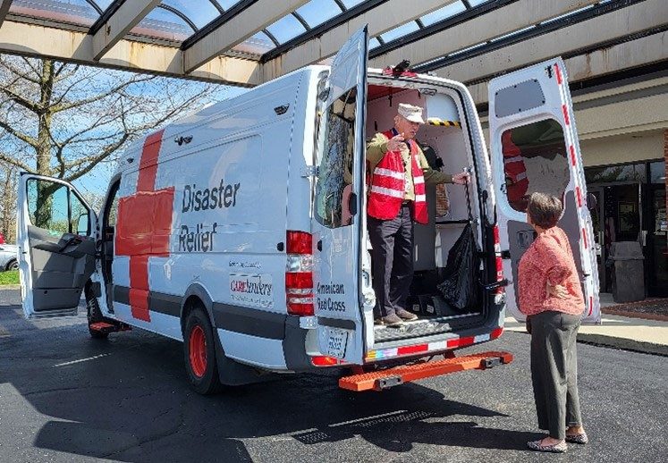 Red Cross volunteer standing in back of Red Cross van with doors open and speaking to person standing on street.