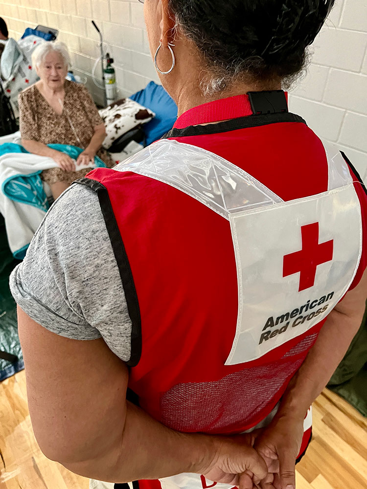 Peggy Mohr sitting on cot in an American Red Cross shelter and the back of Red Cross volunteer Allean Harrington standing by Peggy.