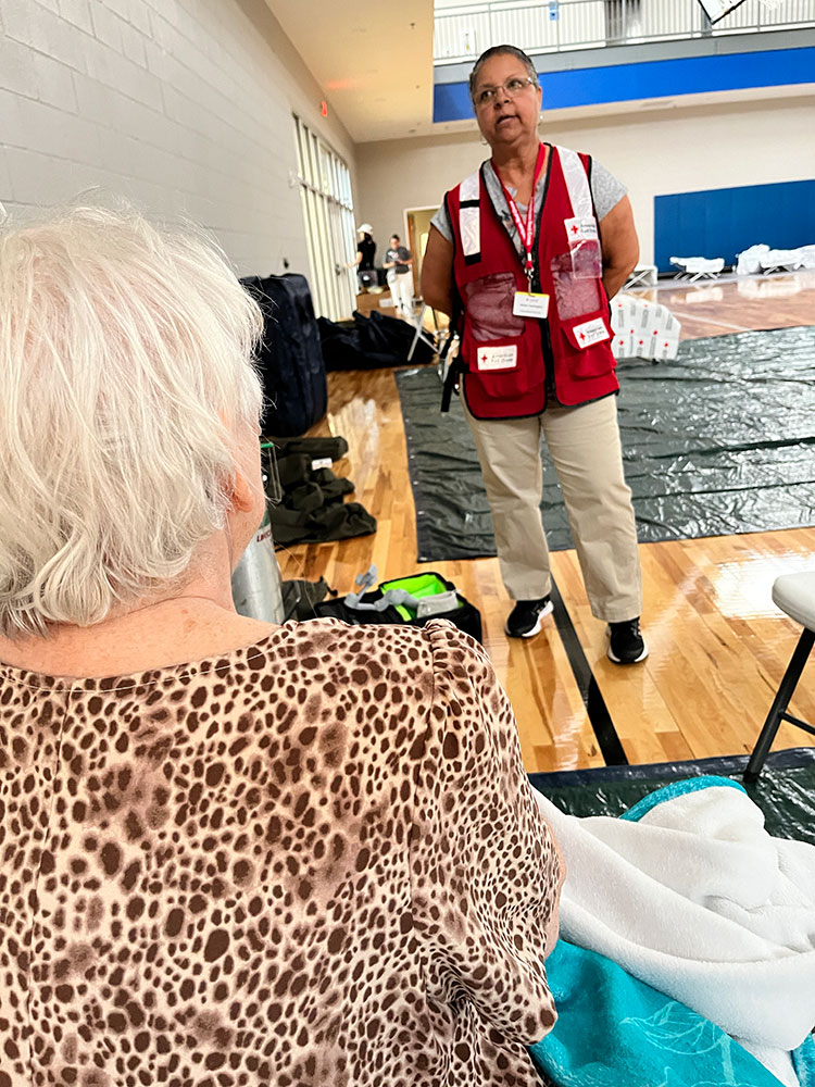 Red Cross volunteer Allean Harrington standing and the back of Peggy Mohr sitting on cot in an American Red Cross shelter.