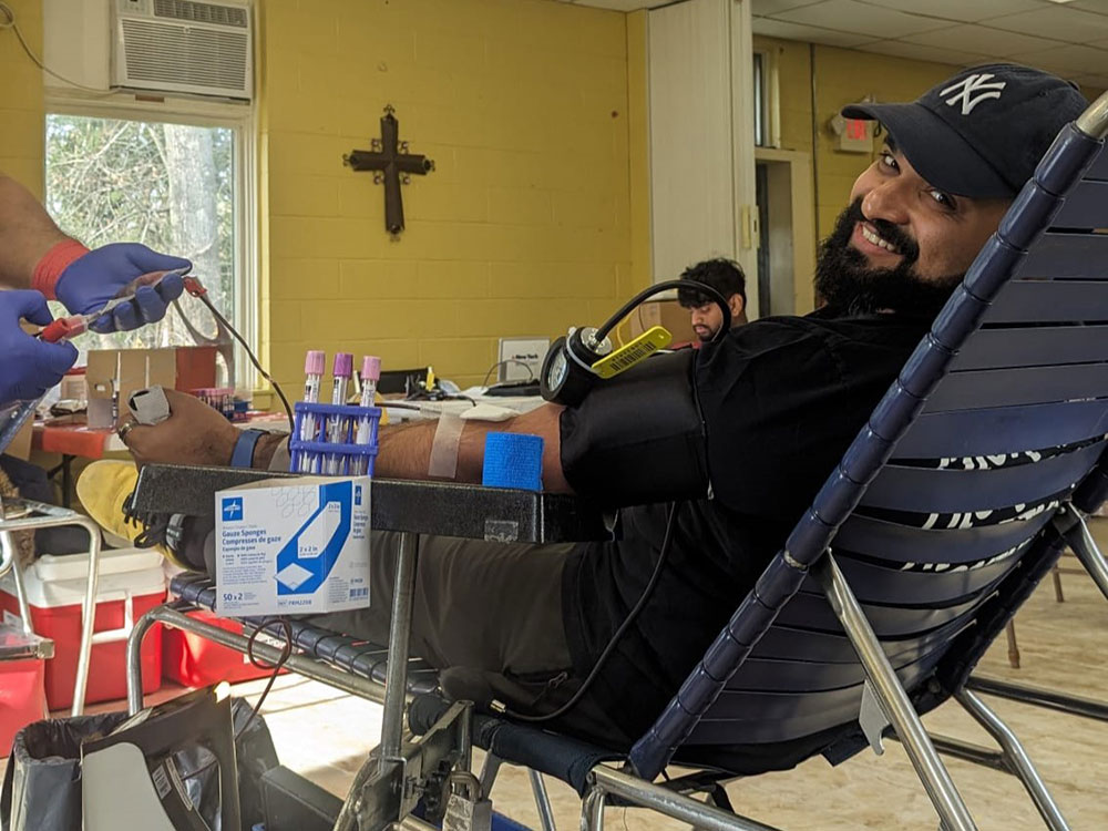 Man wearing a hat, sitting in cot, donating blood and smiling at camera.