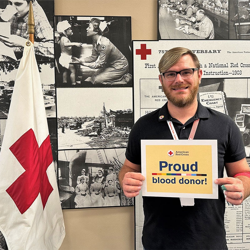 Trevor Jones holding Proud blood donor sign next to Red Cross flag.