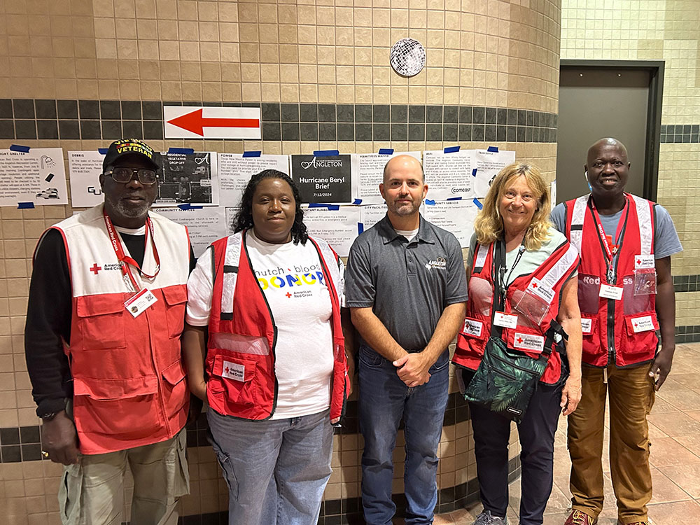 Group of five Red Cross volunteers, wearing Red Cross vests, standing in front of a wall with a sign.