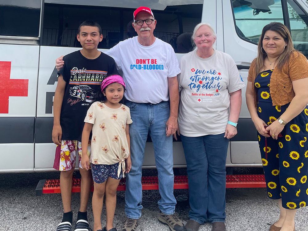 Two Red Cross volunteers stands together with a family smiling in front of a Red Cross vehicle.