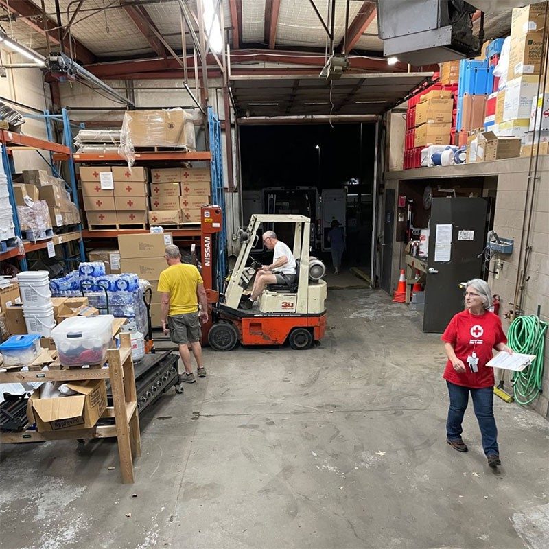 Red Cross disaster workers in warehouse using forklift loading supplies.