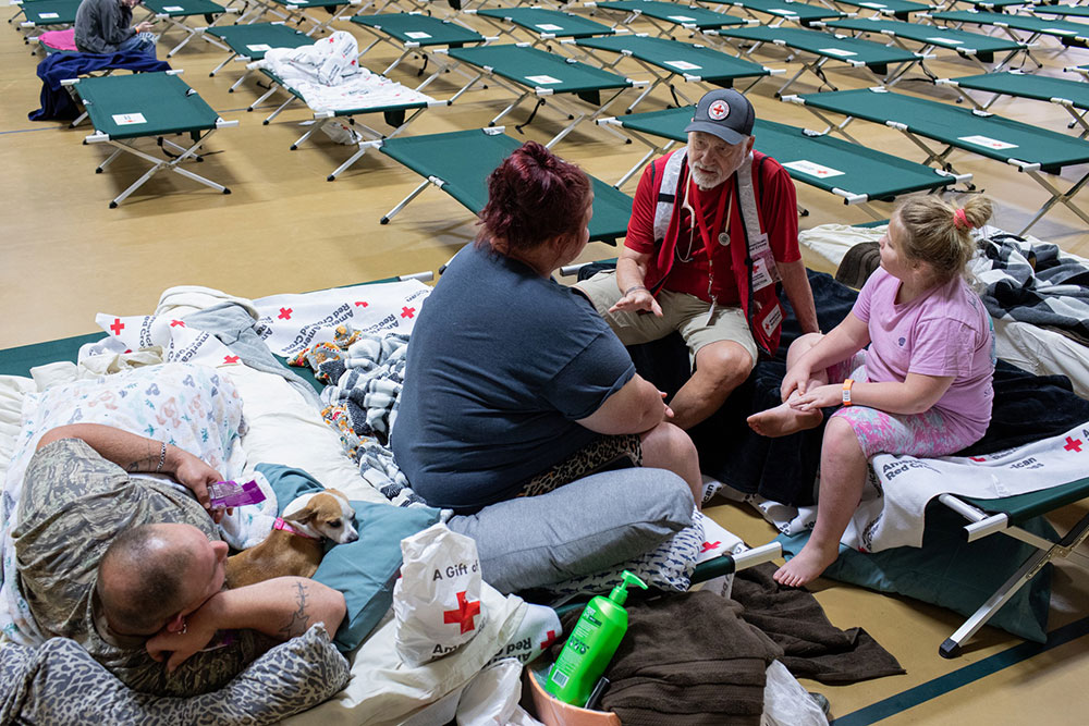 American Red Cross volunteer speaking with family sitting in cots in a disaster shelter.