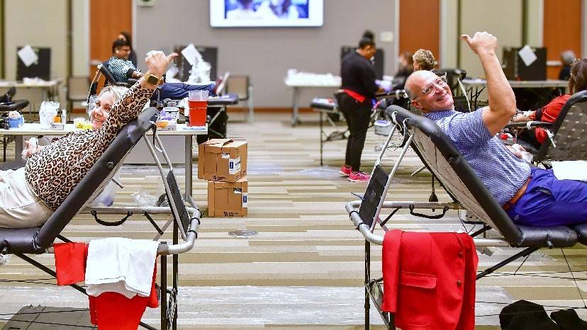 Man and woman sitting in cots donating blood while giving a thumbs up sign.