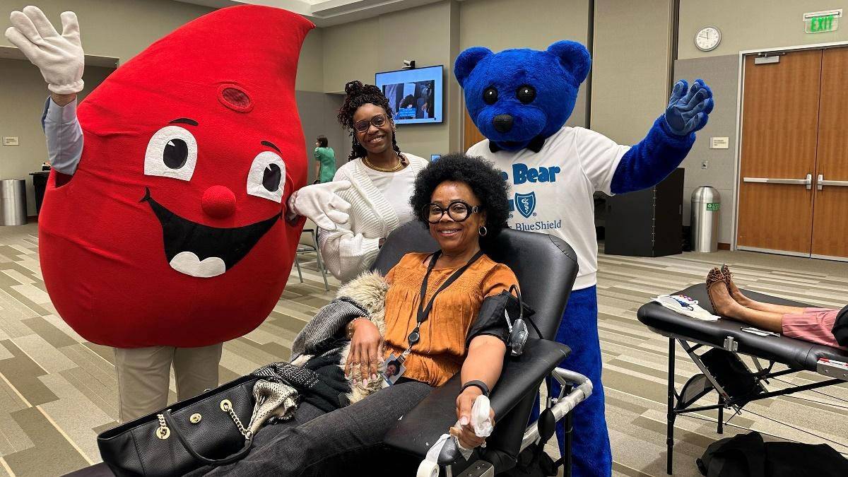 Woman giving blood sitting in cot with 2 mascots and woman standing behind her