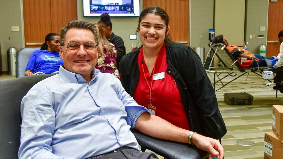 Man giving blood sitting in cot with Red Cross nurse standing behind him