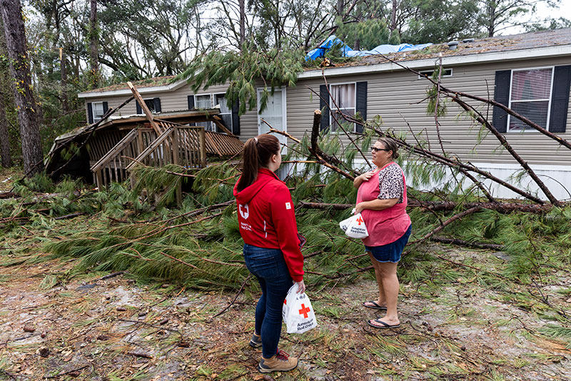 Red Cross volunteer speaking with woman next to damaged house that has fallen trees on it.