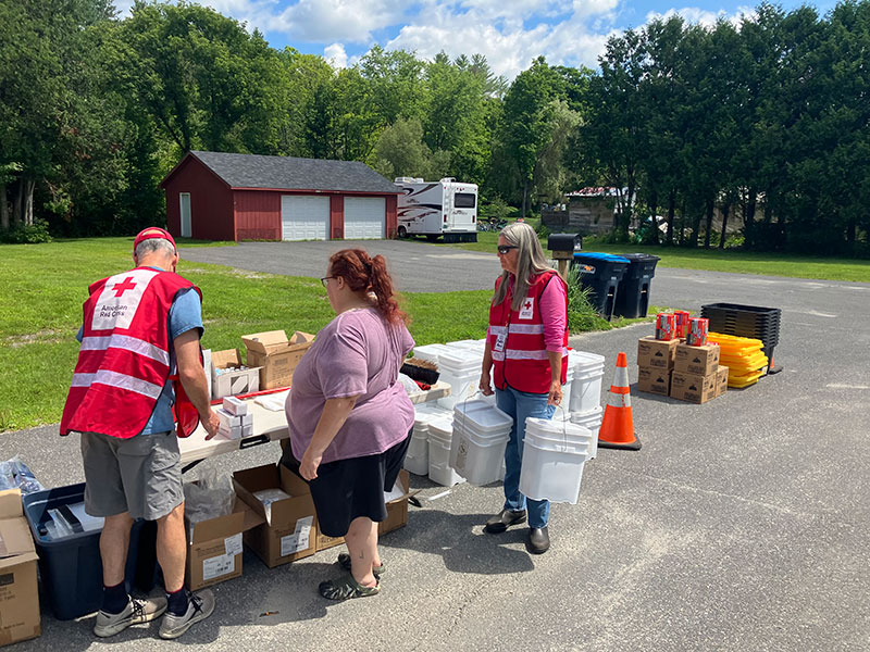 American Red Cross volunteers helping person that's next to a table oustide on street full of supplies.