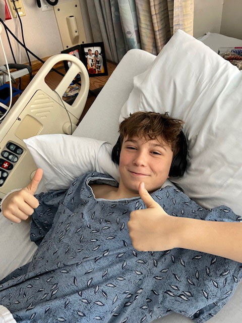 Avery MacNair lying on hospital bed, giving thumbs up and smiling