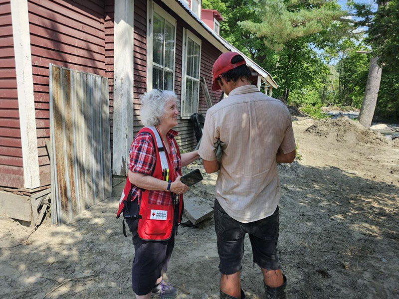 American Red Cross volunteer speaking with person outside house with yard full of dirt.