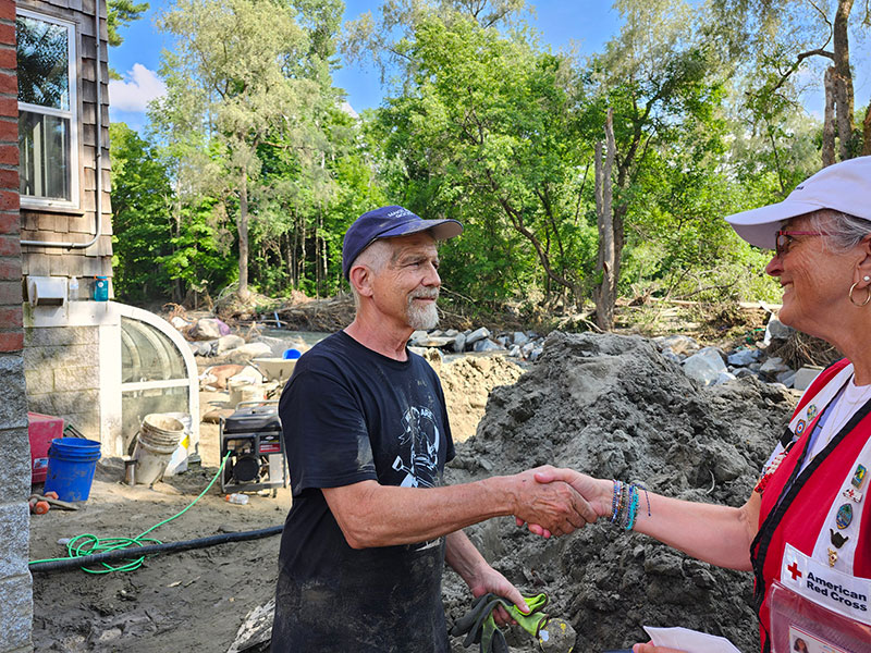 Person shaking hands with American Red Cross volunteer in yard with mounds of dirt, trash and trees in the background.