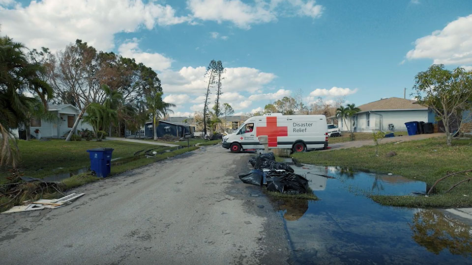 Flood waters next to road in residential area with Red Cross Disaster Relief vehicle parked in a driveway.