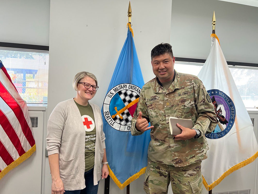 American Red Cross employee and military service member in military outfit with a military flag behind them.