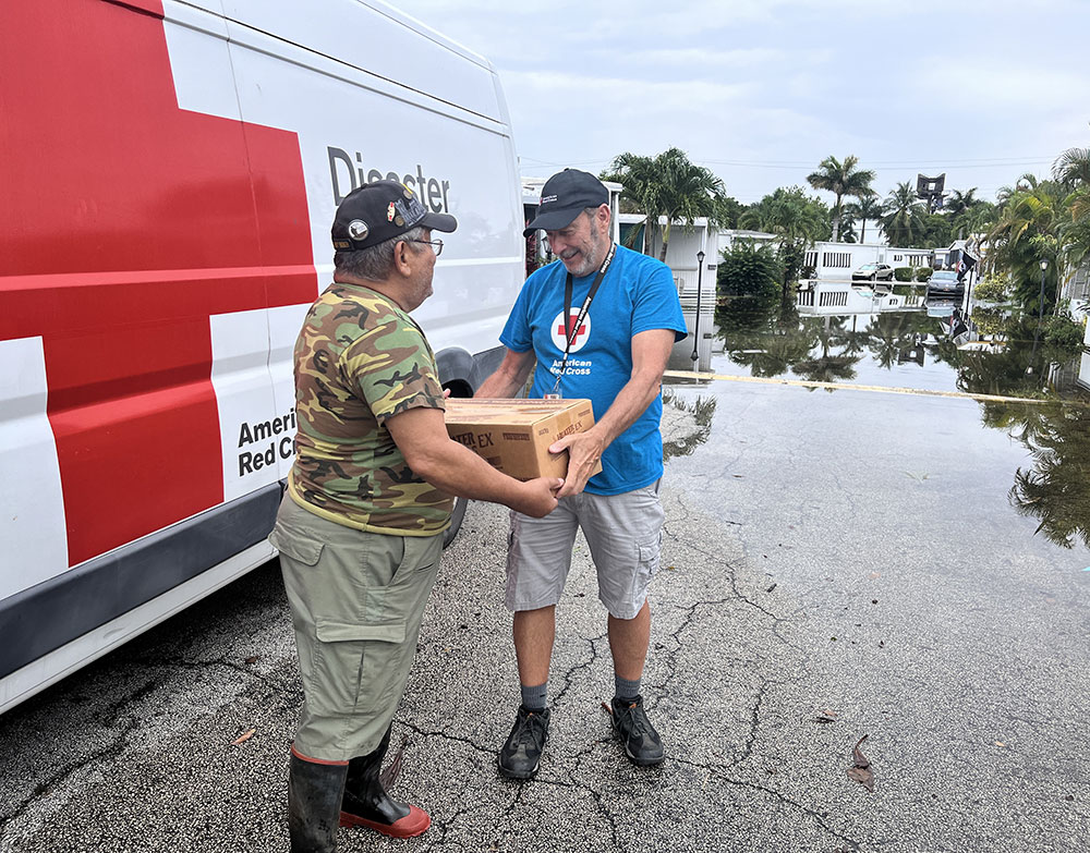 Red Cross volunteers standing next to a Red Cross vehicle handing a box to each other. Mobile home park street flooded in the background.