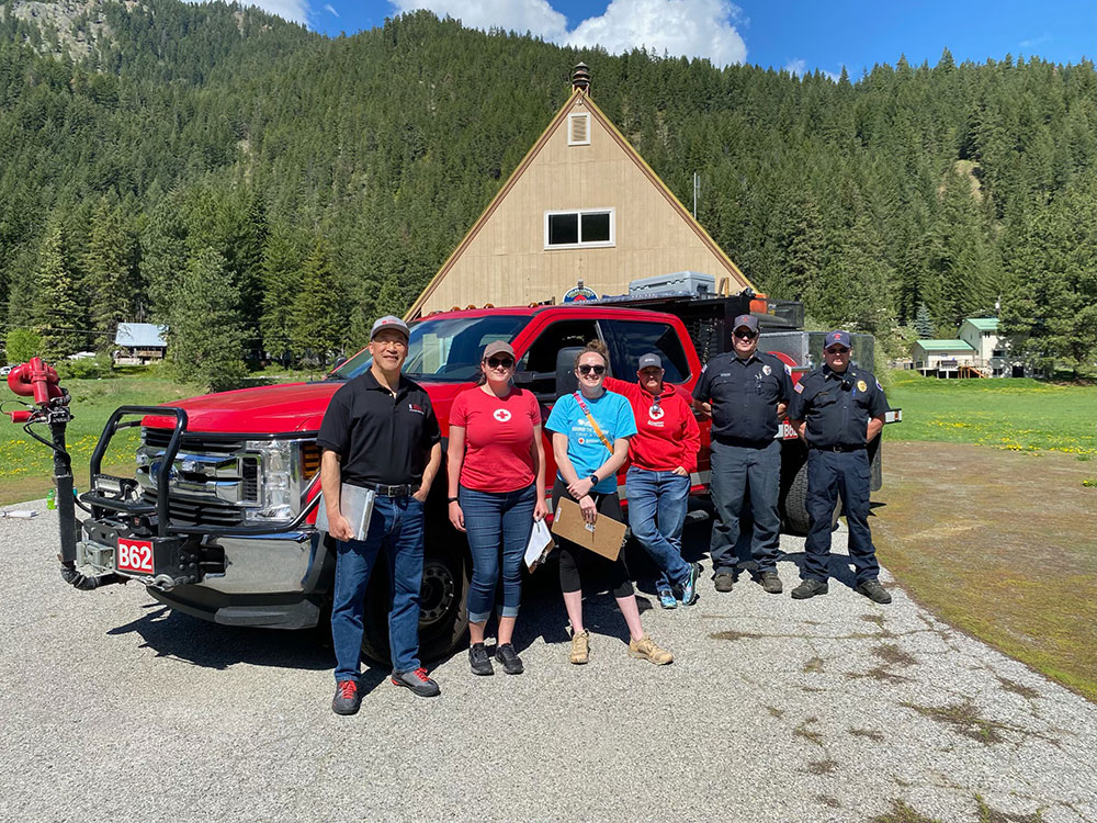 Group of Red Cross volunteers and Firefighters in front of red truck with houses and forest in the background.