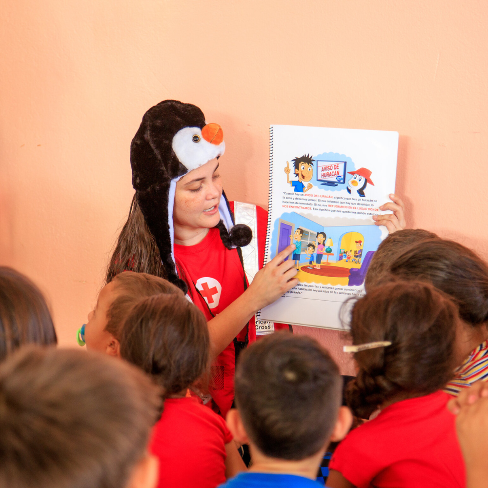 Voluntaria juvenil con sombrero de pingüino, brinda taller de preparación para niños llamado Prepárate con Pedro.