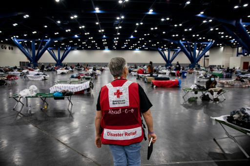 Red Cross Volunteer stands with his back to the camera, facing a room of cots in a Red Cross shelter. 