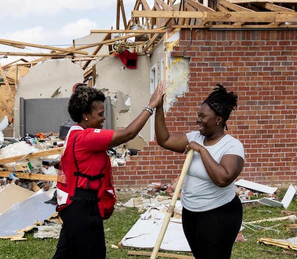 A Red Cross volunteer high fives a woman standing in front of a storm damanged house as they work to clean it.