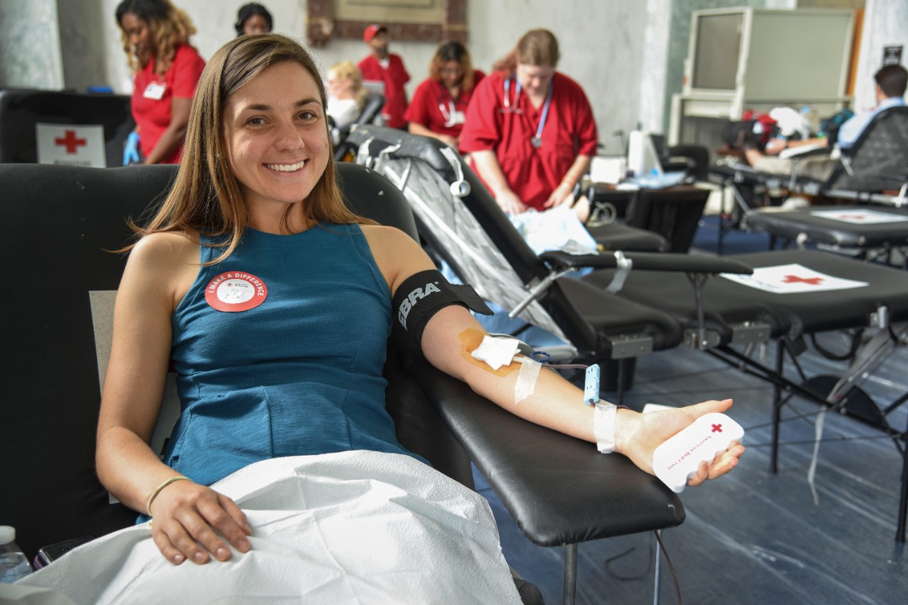 Person sitting in cot donating blood and smiling with American Red Cross nurses in the background.